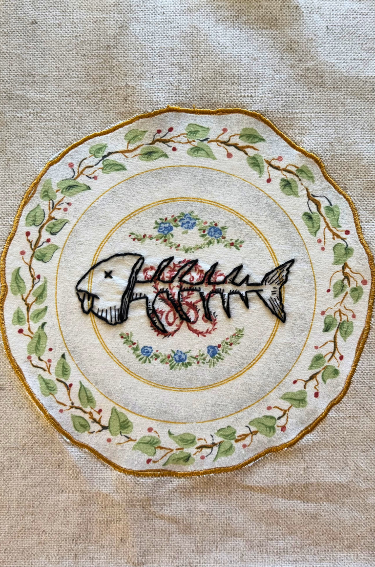 Embroidered fish skeleton on a traditional dinner plate. The fish’s head is still entact— it has x’s for eyes and its tongue is lolling out.